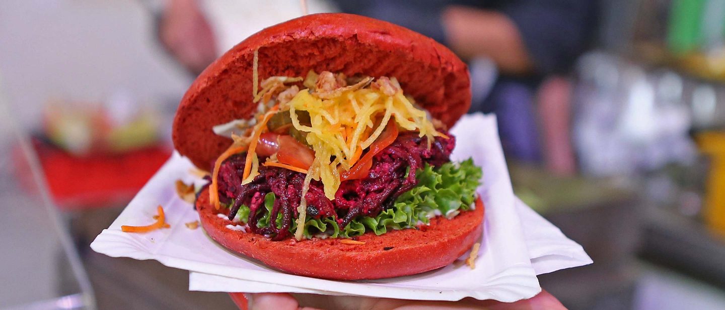 Red burger bun with lettuce, shredded beetroot patty, pickles, and crispy fried topping on a white paper plate.