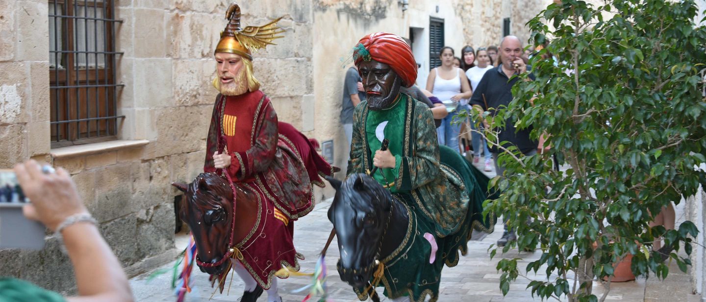Two people in traditional costumes with masks riding horse puppets during a street festival parade.