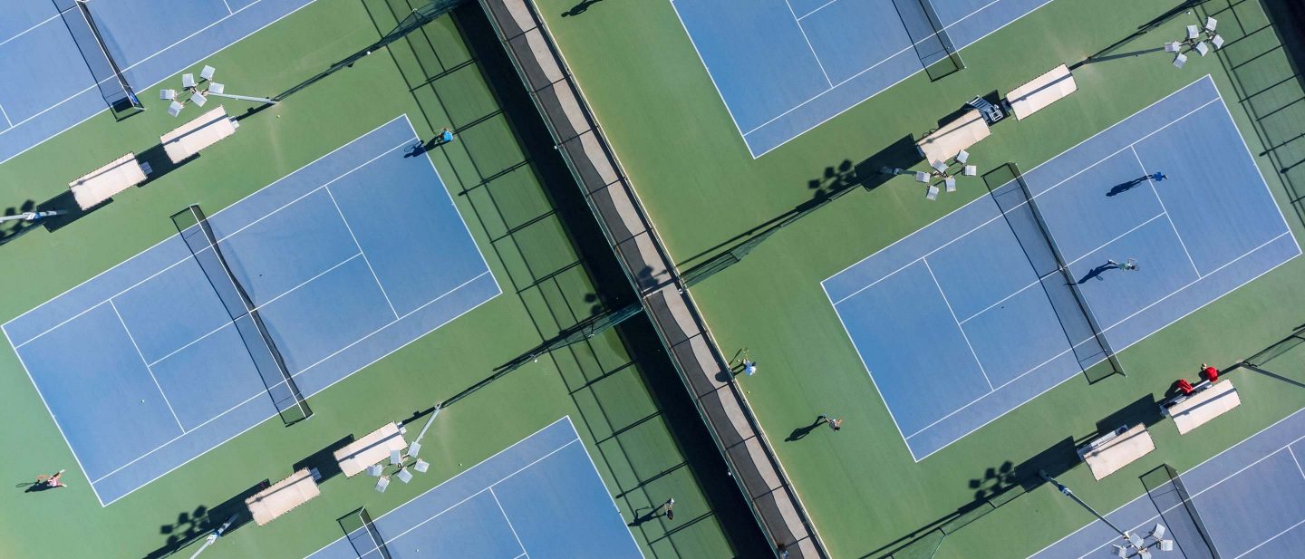 Aerial view of blue and green tennis courts with players and benches under bright sunlight.