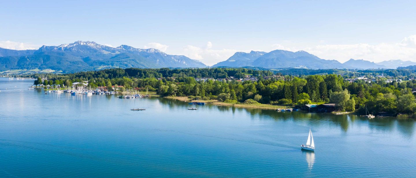 A sailboat on a calm lake with a marina, lush green trees, and distant snow-capped mountains under a clear sky.