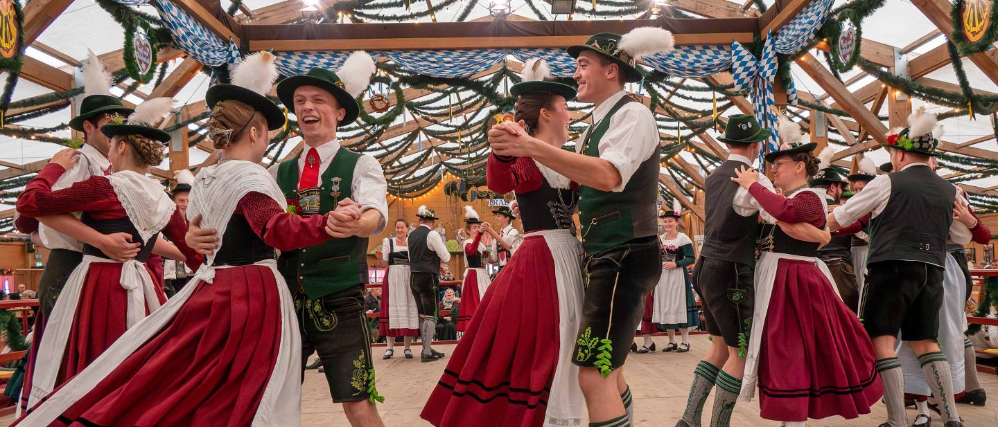 Couples in traditional Bavarian clothing dancing at a festive event inside a decorated wooden hall.