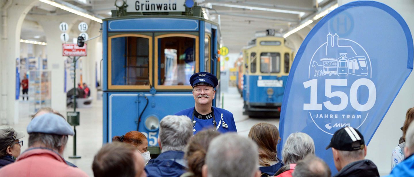 Man in vintage uniform speaking to crowd in front of historic blue trams at tram museum event.