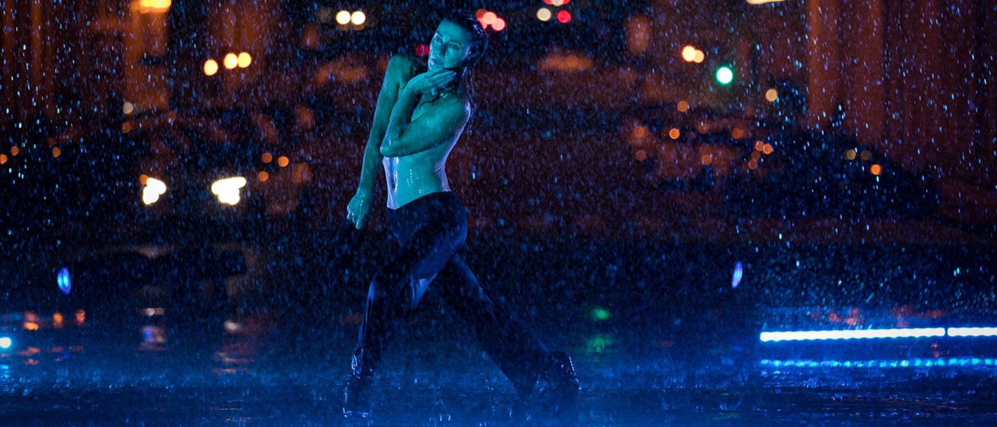 Woman dancing in the rain at night with city lights blurred in the background and blue lighting on the ground.