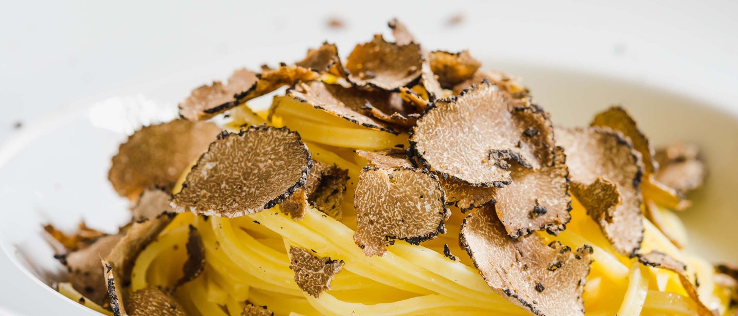 Spaghetti pasta topped with thinly sliced black truffles served in a white bowl.