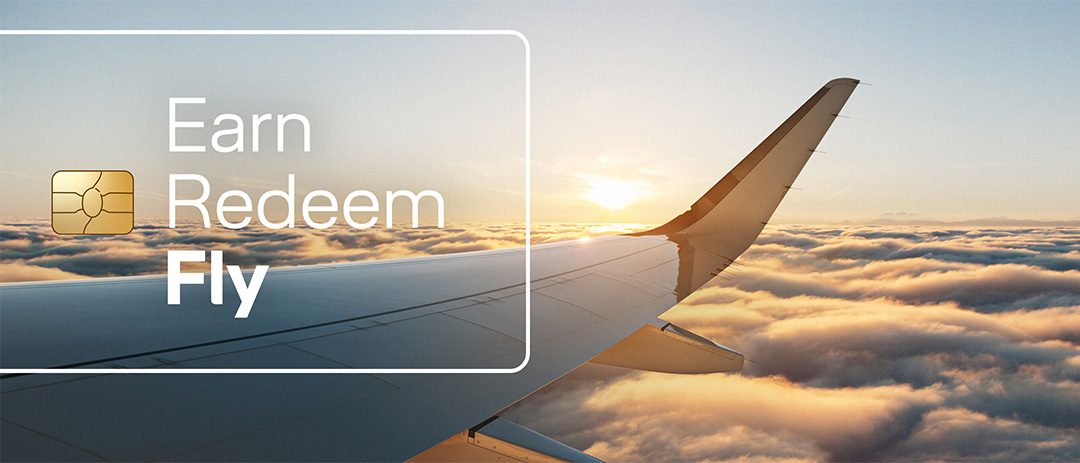 Airplane wing rises above golden clouds at sunset, with “Earn, Redeem, Fly” text and a gold card icon, evoking aspirational travel rewards.
