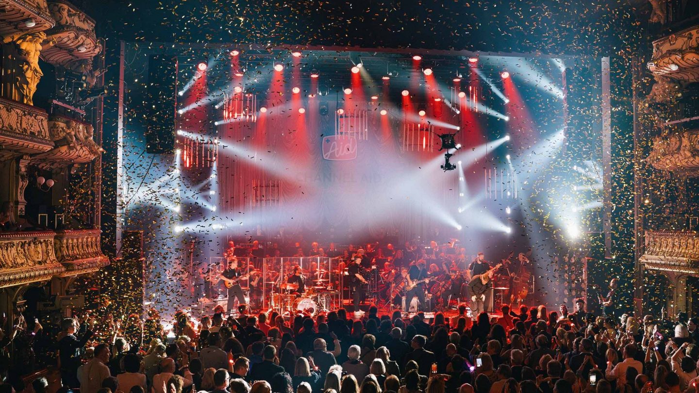 A concert in a glamorous theater hall with a band, orchestra, spotlights, and confetti