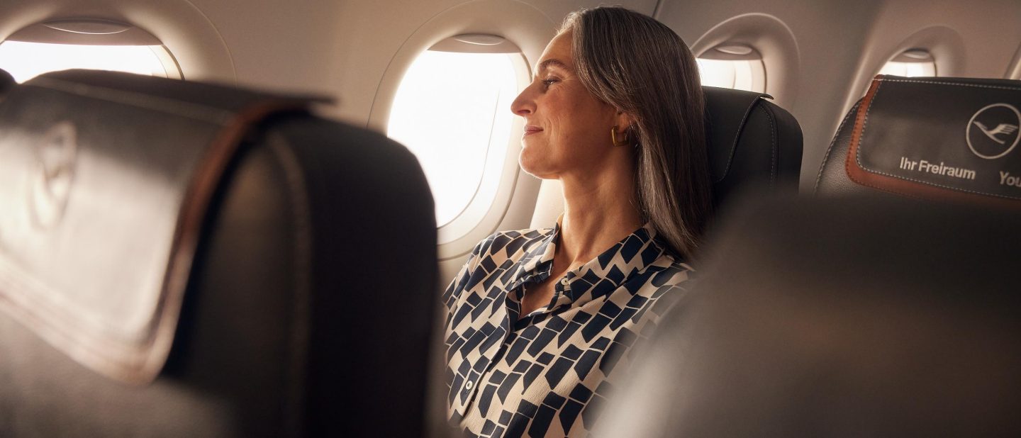 Woman seated by an airplane window, eyes closed and enjoying the sunlight; blurred seats frame her calm, relaxed journey.